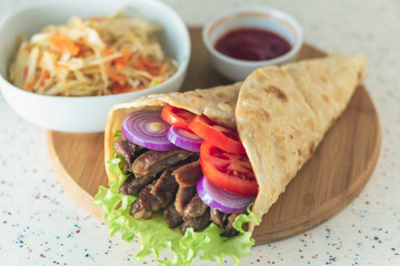 Handmade doner kebab is lying on the white table made of artificial stone Shawarma on the wooden cutting board with chicken meat, onions, salad lies on a dark old wooden table.