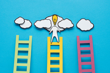 cardboard man with light bulb head climbing one of three ladders on blue background, ideas concept