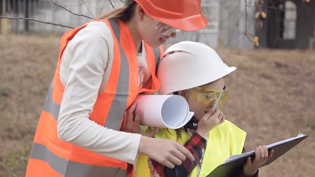 Two children girls in construction helmets looking at white sheet of paper or drawing and smiling