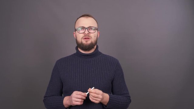Portrait of a young bearded man in glasses trying to quit smoking. Isolated on grey background.
