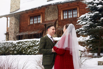 Cute stylish couple on wedding day. Bride and groom meet for the first time. First look. Winter wedding on snowfall with nice wooden cottage chalet on ski resort. Woman wearing knitted cardigan