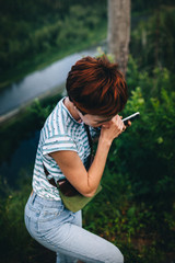 Red-haired hipster girl with a phone, stands against the background of the river.