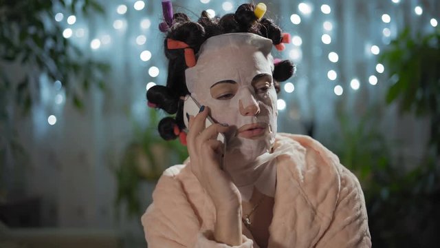 Close-Up, Beautiful Woman in a Rejuvenating Mask with Curlers Talking on the Phone Sitting in the Room
