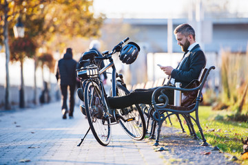 Businessman commuter with bicycle sitting on bench in city, using smartphone.