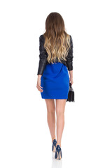 Walking Woman In Blue Mini Dress, High Heels And Leather Jacket. Rear View.