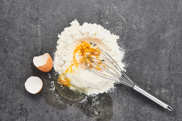 Mixing egg and flour. Egg, flour and whisk on dark grey concrete background. Ingredients for dough...