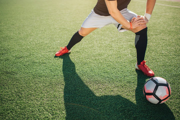 Football player stand on green lawn and stretch legs. He leans to one of it. Sun is shining...
