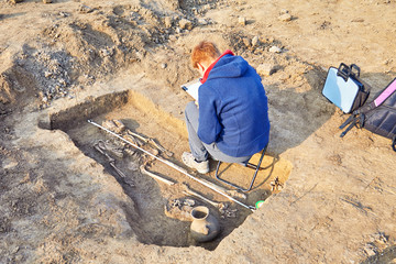 Real archaeological excavation. Archaeologist in a digger process, draws the tomb with human bones, part of skeleton and skull in the ground. Outdoors