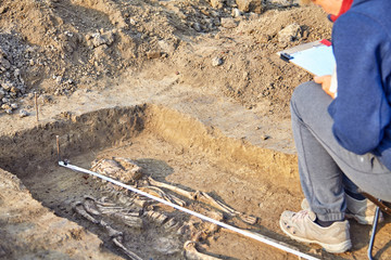 Real archaeological excavation. Archaeologist draws the tomb with human bones, part of skeleton and skull in the ground. Outdoors, copy space, close up.