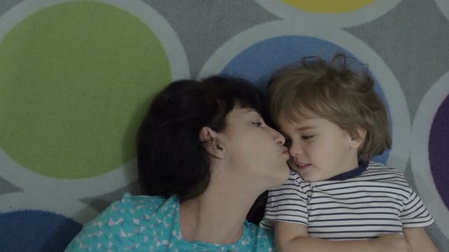 Excited young mother playing lying on the floor and taking a selfie. Mother and son monkeying around on camera and kissing. Portrait of mom and baby closeup. Slow Motion.