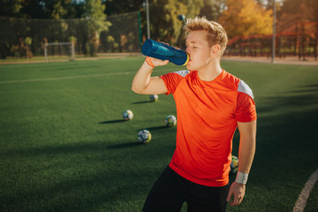 Young athlete stand on lawn outside and drink water from bottle. He has rest. Four balls are behind...