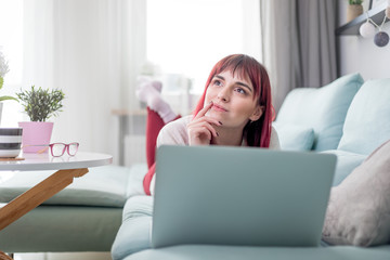 Fototapeta premium Smiling woman lying on sofa and using laptop while thinking and planning future at home