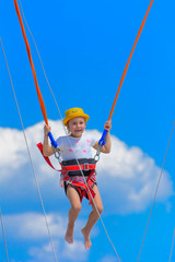 A little girl jumps high on a trampoline with rubber ropes against the blue sky and white clouds. Adventures and extreme sports. Concept of summer vacation, jumping.