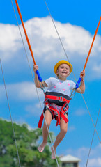 A little girl jumps high on a trampoline with rubber ropes against the blue sky and white clouds. Adventures and extreme sports. Concept of summer vacation, jumping.
