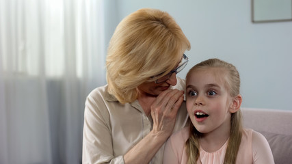 Positive blond grandmother whispering secret to surprised granddaughter, family