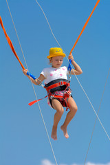 A little girl jumps high on a trampoline with rubber ropes against the blue sky and white clouds. Adventures and extreme sports. Concept of summer vacation, jumping.