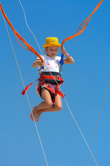 A little girl jumps high on a trampoline with rubber ropes against the blue sky and white clouds. Adventures and extreme sports. Concept of summer vacation, jumping.