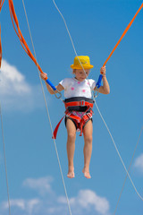 A little girl jumps high on a trampoline with rubber ropes against the blue sky and white clouds. Adventures and extreme sports. Concept of summer vacation, jumping.