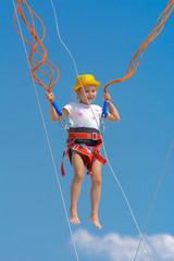 A little girl jumps high on a trampoline with rubber ropes against the blue sky and white clouds. Adventures and extreme sports. Concept of summer vacation, jumping.