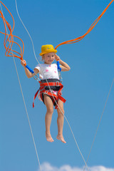 A little girl jumps high on a trampoline with rubber ropes against the blue sky and white clouds. Adventures and extreme sports. Concept of summer vacation, jumping.