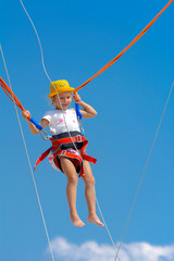 A little girl jumps high on a trampoline with rubber ropes against the blue sky and white clouds. Adventures and extreme sports. Concept of summer vacation, jumping.