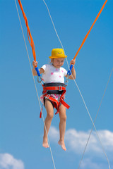 A little girl jumps high on a trampoline with rubber ropes against the blue sky and white clouds. Adventures and extreme sports. Concept of summer vacation, jumping.