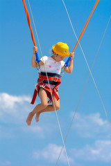 A little girl jumps high on a trampoline with rubber ropes against the blue sky and white clouds. Adventures and extreme sports. Concept of summer vacation, jumping.