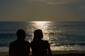 Silhouettes of a couple, man and woman are sitting on a sandy beach and looking at the sunset on the sea on a vacation. Copy space.    