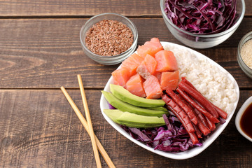 poke bowl Hawaiian food. a plate of rice, salmon, avocado, cabbage and cheese, next to sesame and soy sauce. on a brown wooden table.