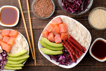 poke bowl Hawaiian food. a plate of rice, salmon, avocado, cabbage and cheese, next to sesame and soy sauce. on a brown wooden table. top view