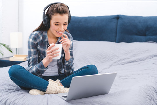 happy young woman in headphones holding cup and using laptop on bed