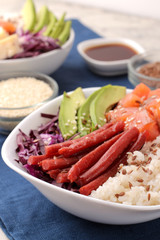 poke bowl Hawaiian food. a plate of rice, salmon, avocado, cabbage and cheese. near skunk and soy sauce on a blue napkin on a light background.
