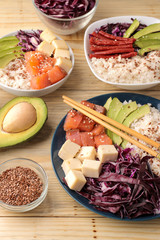 poke bowl Hawaiian food. a plate of rice, salmon, avocado, cabbage and cheese. next to sesame and fresh avocado on a natural wooden table