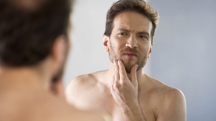 Fototapeta na wymiar Middle-aged man critically looking at his beard in mirror, morning ritual