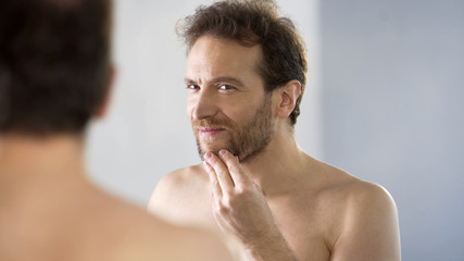 Fototapeta premium Topless male critically looking at his beard in mirror, morning ritual, bathroom