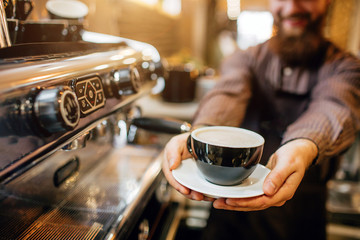 Cut view of young bearded man holding cup of coffee in hands. He stand in kitchen at coffee machine.