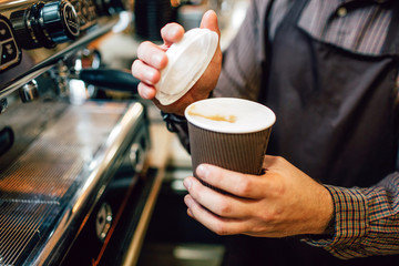 Close up of barista holding cup of coffee and lid. He is going to put lid on cup. Guy stand at coffee machine.