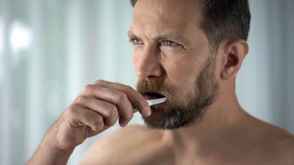 Aging man brushing teeth and looking at mirror, morning procedure, dental care
