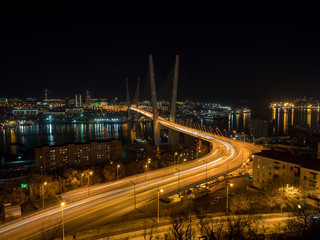 Zolotoy most or Golden Bridge, Vladivostok, Primorsky region, Russia. The lights of the night city under a cable-stayed bridge over the river with construction cranes on the shore. December, 2018