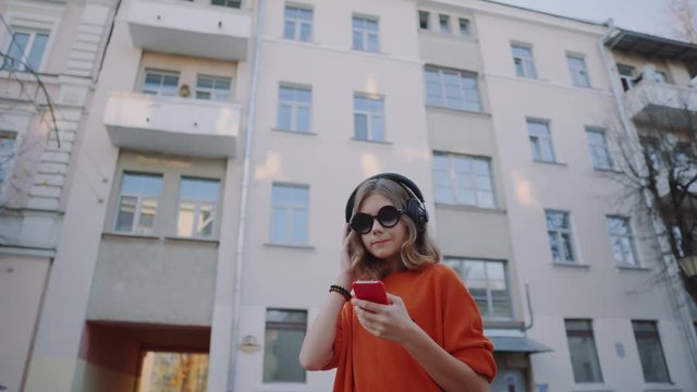 cute young girl listening music in headphones, urban style, stylish hipster teen choosing track on mobilephone and smile infront of oidsity buildings, orange crazy street style