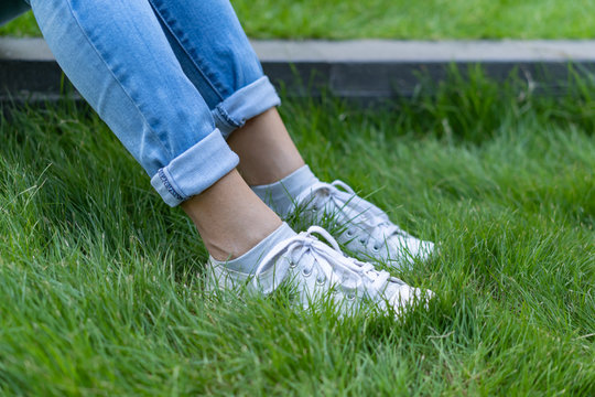 Close Up Legs Of Woman Dressed In Jeans And Sneakers Step On The Grass Of The Garden In A Park.