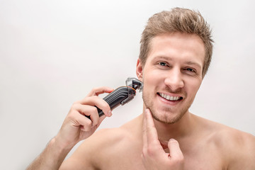 Cheerful young man shaving with machine. He smiles and touches chin. Isolated on white packground.
