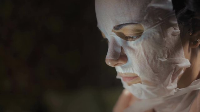 Close-up, Woman in Rejuvenating Mask Looks at Smartphone Screen Sitting in the Dark Room