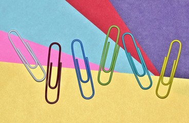 Six scattered colorful paper clips against a bright multi-colored paper background. Paper clips are arranged randomly.