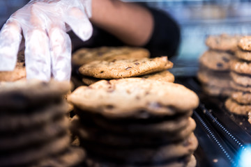 Closeup of chocolate chip cookies on display in bakery pile stack with employee worker baker glove on hand touching baked goods sweets