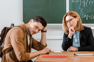 Young student sitting with backpack near female teacher and holding head