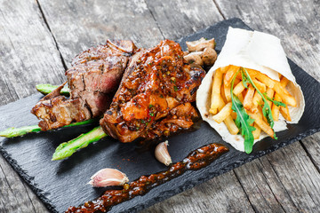 Grilled pork steaks with herbs, asparagus, garlic, mushrooms and potato fry on stone slate background on wooden background close up. Hot Meat Dishes. Top view, flat lay
