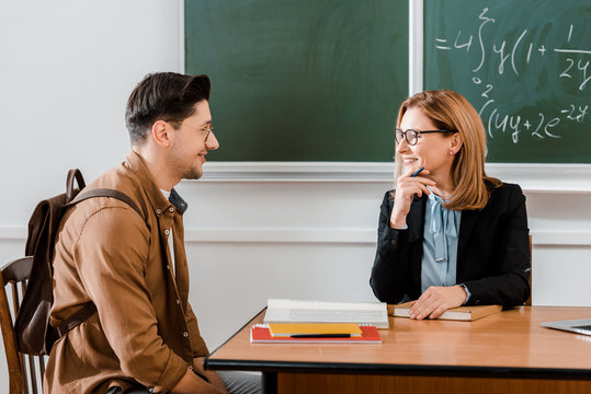 Female teacher smiling near young student with backpack in classroom - Powered by Adobe