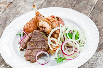 Grilled meat on plate, chicken legs, beef steak and onion on wooden background close up. Top view