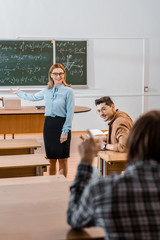 selective focus of smiling female teacher pointing at chalkboard with hand while male student looking at classmate